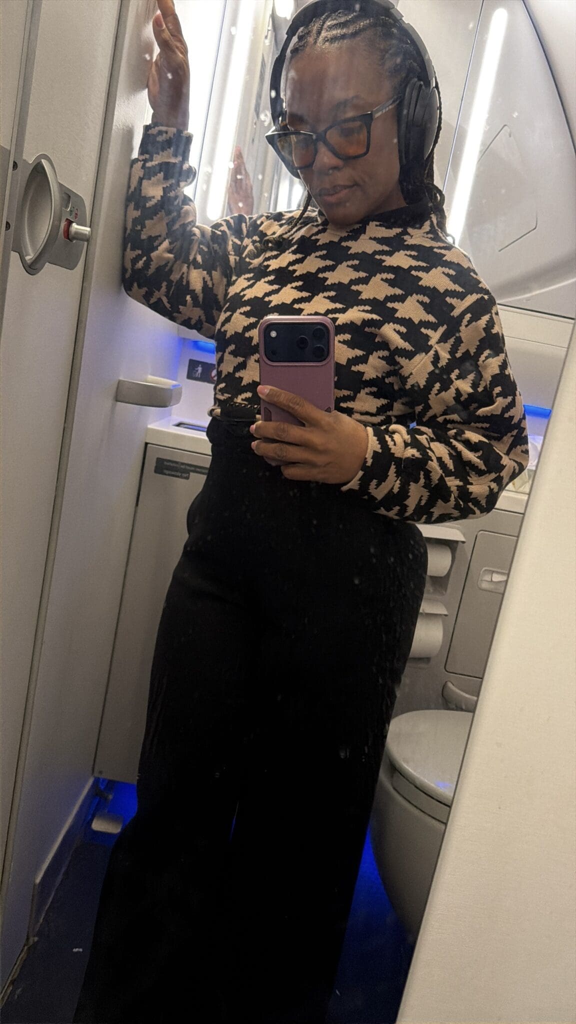 Woman taking an airplane bathroom mirror selfie wearing a stylish black travel outfit, houndstooth sweater, travel sunglasses, and noise-canceling headphones.
